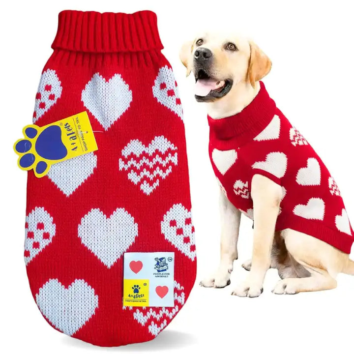 A red knit pet sweater with a white heart pattern is shown both flat on the left and worn by a light-colored dog on the right. The sweater features a ribbed red neck, a red paw print patch, and a white brand tag.