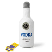 A white plush dog toy shaped like a vodka bottle with a gray cap, featuring a glittery paw print and the word 'VODKA' in blue. A small yellow and purple paw-shaped tag is placed beside it on a white background.