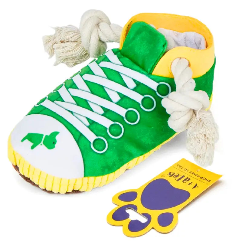 A novelty dog toy shaped like a sneaker, featuring a green canvas upper, white laces, a yellow collar and sole trim, a white toe cap with a green dog silhouette, and white rope laces. A yellow paw-shaped tag is shown next to it on a white background.
