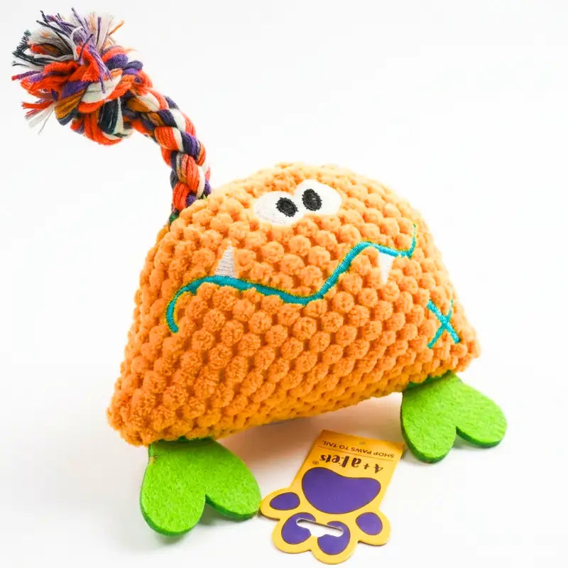 An orange textured monster dog toy with green felt feet, embroidered facial features, and a multicolor braided rope tail, shown on a white background with a small tag.
