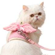 A white long-haired cat wearing a pink pet harness with angel wings and a matching pink leash, photographed against a plain white background.