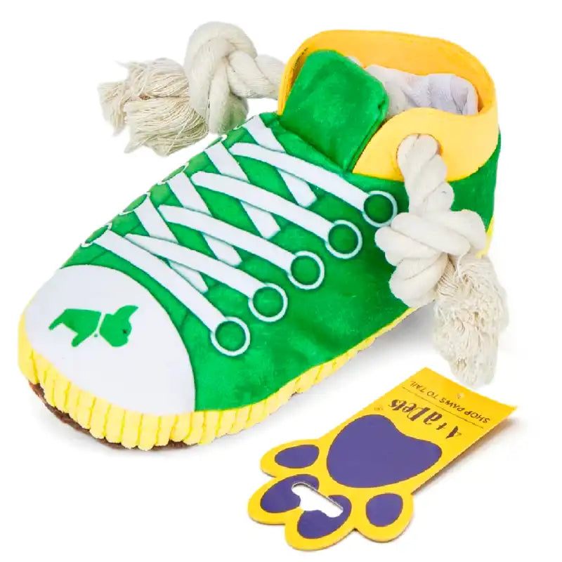 A novelty dog toy shaped like a sneaker, featuring a green canvas upper, white laces, a yellow collar and sole trim, a white toe cap with a green dog silhouette, and white rope laces. A yellow paw-shaped tag is shown next to it on a white background.