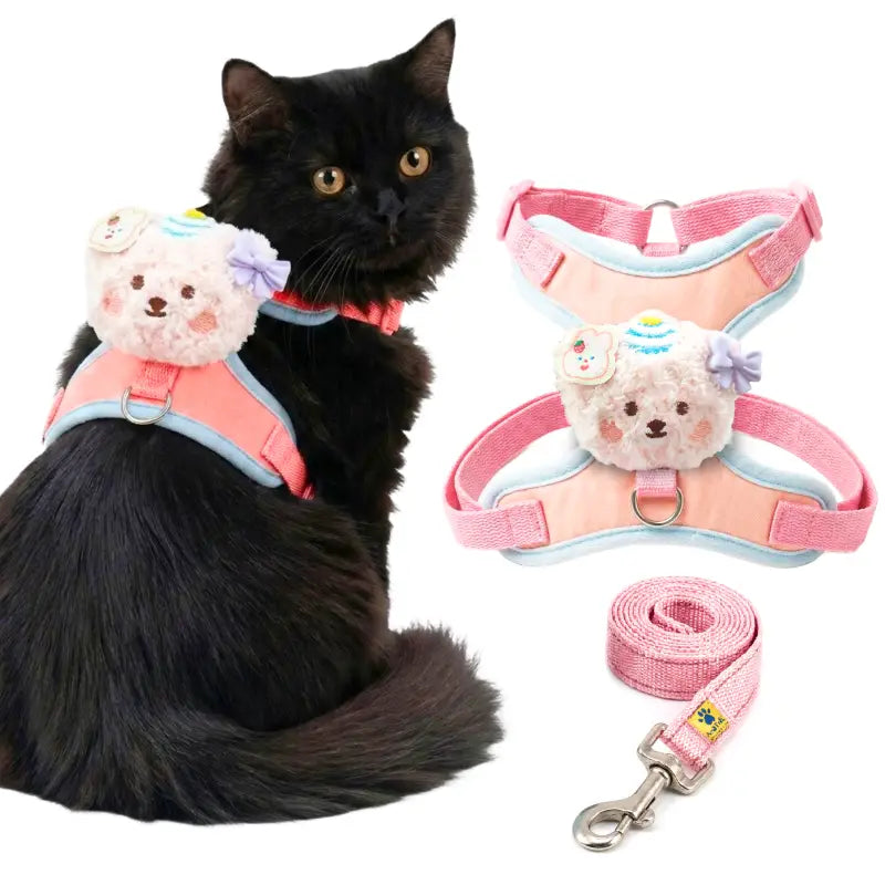 A black cat wearing a pink and light blue pet harness with a plush teddy bear face accessory, shown alongside the same harness and matching leash on a white background.