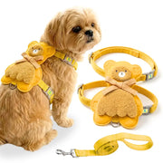 A small fluffy dog wearing a yellow pet harness with a teddy bear applique, shown beside the harness and matching leash on a white background.
