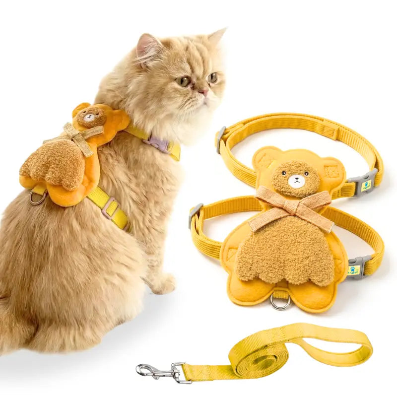 A fluffy cream-colored cat wearing a yellow harness with a plush teddy bear attachment, shown next to the harness laid flat and a matching yellow leash on a white background.