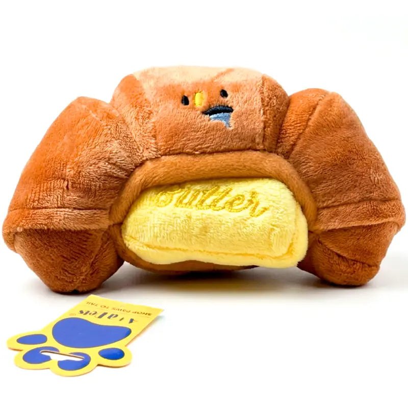 A plush croissant toy with a yellow butter insert and embroidered face, shown on a white background with a small yellow and blue tag in front.