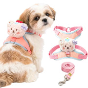 A small fluffy dog wearing a pink and light blue harness with a plush bear face accessory, shown beside the harness laid flat and a matching pink leash on a white background.
