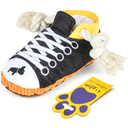 A plush dog toy shaped like a sneaker, with a black upper, white laces, white rubberized toe cap, orange sole, and white rope handles. A yellow and purple paw-shaped tag is placed next to it on a white background.