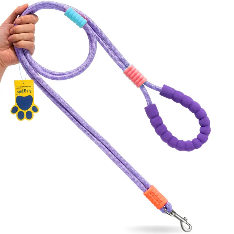 A purple braided dog leash with purple rubber handle grips, orange and pink foam accents, and a yellow paw-shaped tag, held by a hand on a white background.