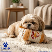 A small cream-colored Shih Tzu dog lies on a white textured rug in a cozy living room, holding a plush novelty toy shaped like a coffee cup with orange graphics.