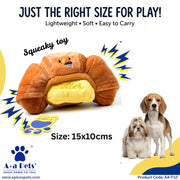 A plush dog toy shaped like a croissant with a yellow butter filling, shown on a white background with text reading 'JUST THE RIGHT SIZE FOR PLAY!' and 'Squeaky toy.' Two small dogs are pictured at the lower right.