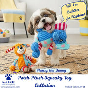 A small fluffy dog stands on a patterned rug holding a blue crocheted elephant squeaky toy, with an orange crocheted bunny toy beside it. The scene is indoors with furniture blurred in the background.